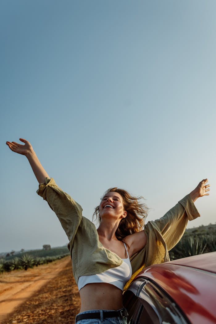 Woman joyfully stretches arms beside car on a sunny rural road, embracing freedom.